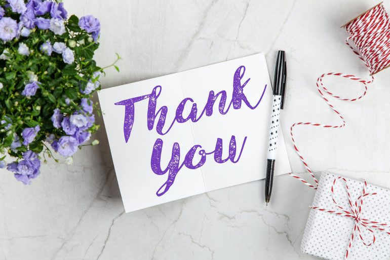 Papel com escrito "Thank You" em cima de uma superfície, com flores e caneta ao lado. Indica gratidão. Créditos: Pexels.